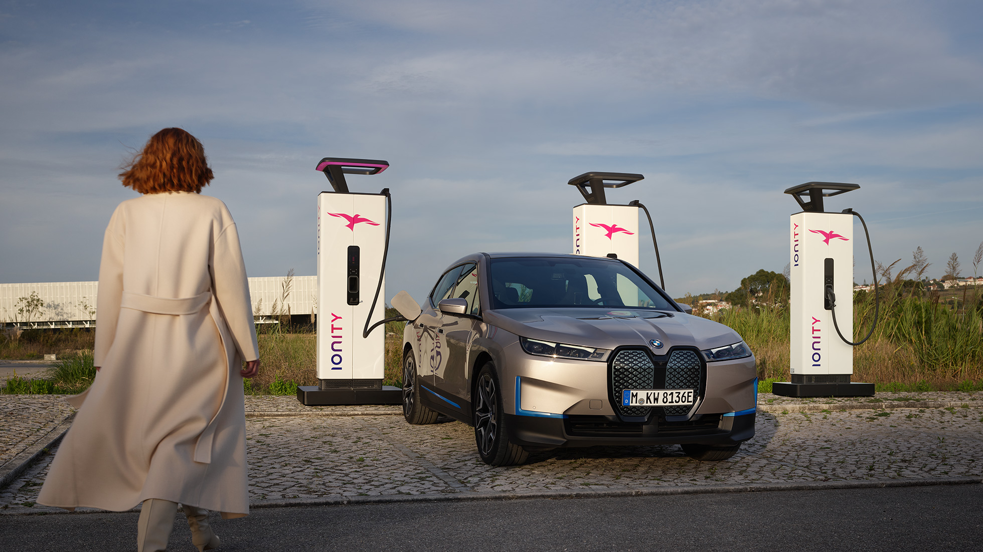 Three quarter front view BMW iX i20 2020 metallic at a charging station BMW iX i20 2020 metallic 3/4 front view at a charging station with woman walking