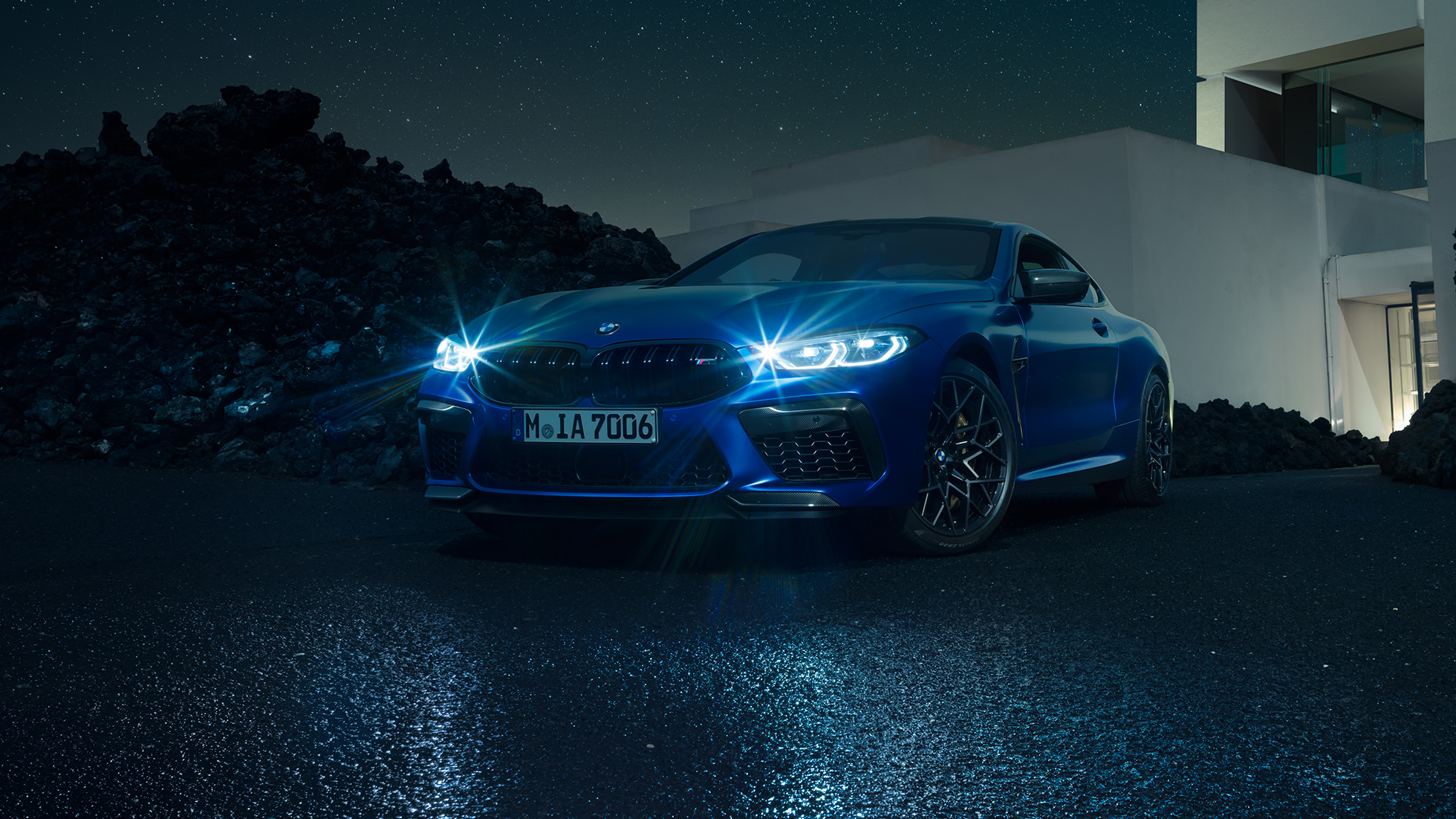 BMW M8 Competition Coupé in Marina Bay Blue metallic with shining LED headlights, standing three-quarter front view in front of villa at night.